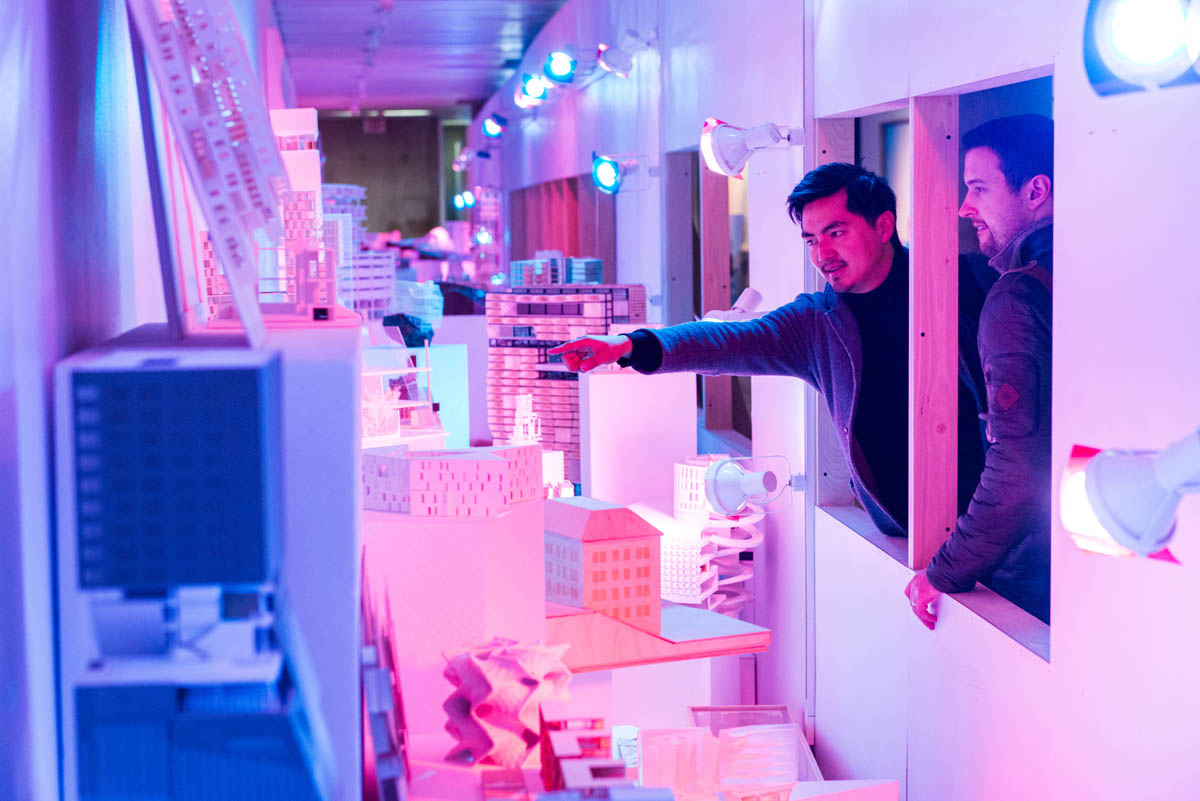 Two people point and lean through the square windows of a plain plywood wall obstructing many student models brightly cross-lit in blue and pink hues.