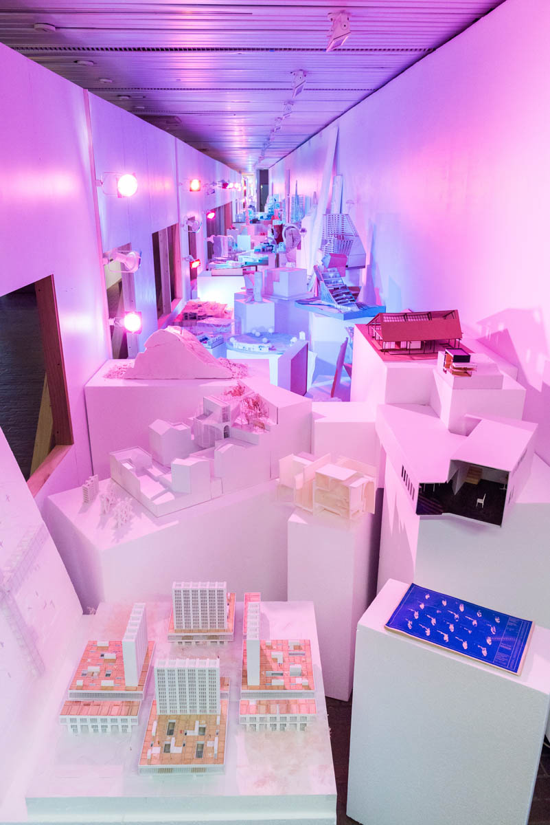 A long view of behind a plain plywood wall obstructing many student models lit brightly in blue and pink hues.