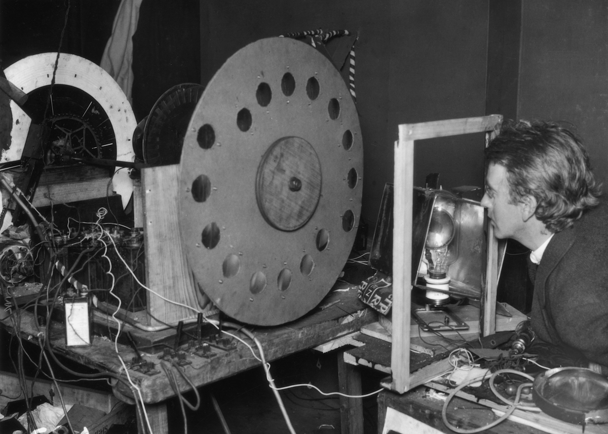 A white man, John Logie Baird, is looking into his mechanical television which is made of a series of wires and moving circles.