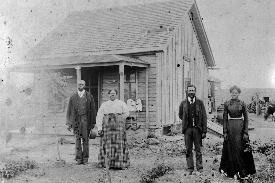 The image is black and white. Two couples stand outside of a small wood-paneled building. Both men are wearing dark-colored suits and both women are wearing long skirts, one plaid and one black, and long sleeved tops, one white and one black.