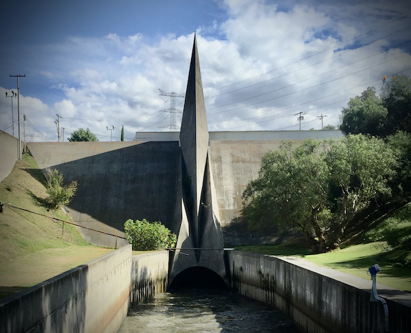 A small waterway enters a tunnel below a large spire, surrounded by sparse trees and a blue sky with clouds above.