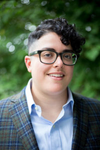 Aimi Hamraie, an olive-skinned west Asian non-binary person with short, dark curly hair and rectangular glasses smiles slightly at the camera. They wear a blue shirt, blue and green plaid blazer, and stand in front of a blurred green background.