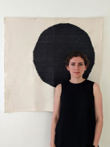 Headshot of Yasmin Vobis, dressed in all black and standing in from of a painted black circle on canvas.
