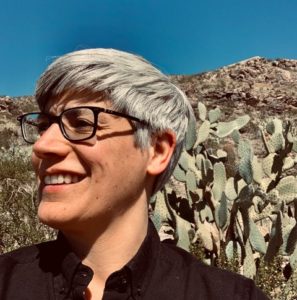 Headshot of Kathryn Yusoff, who wears glasses and a black shirt and stands in front of a hillside with cactus.