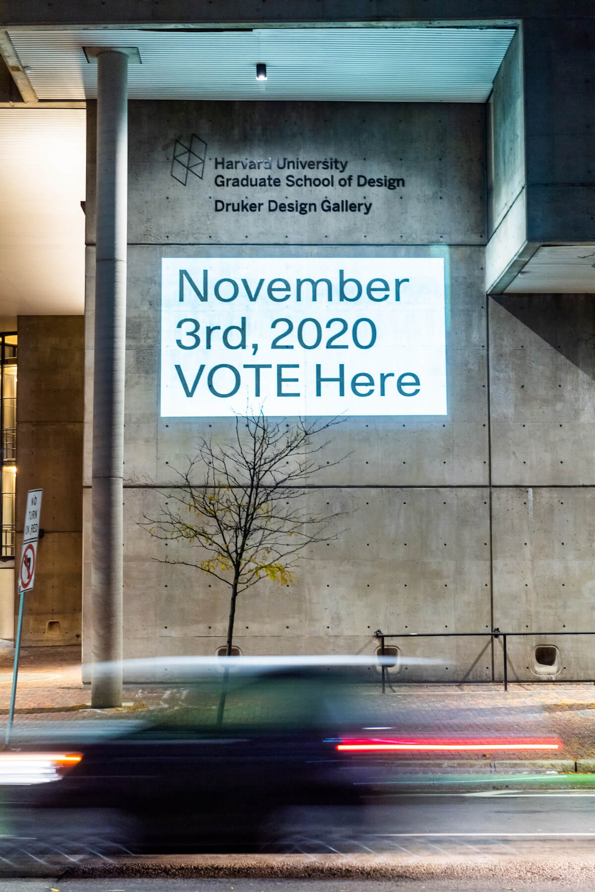 Gund Hall at night with projection reading "November 3rd, 2020 VOTE Here" illuminated on the side of the building.