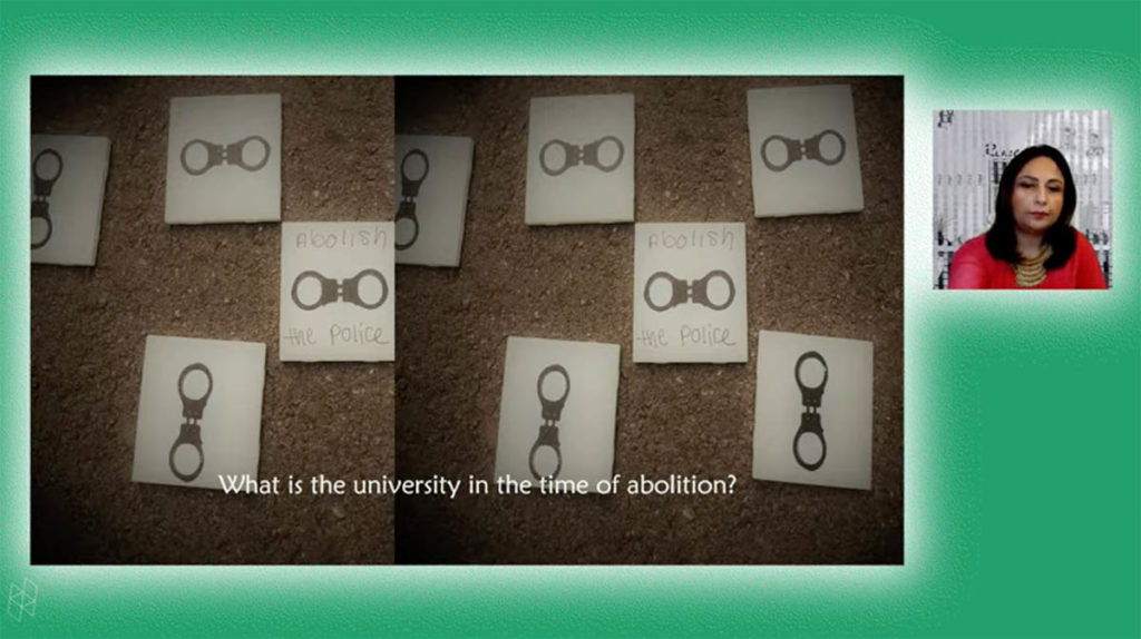 Screenshot from a virtual event. Ananya Roy, who has long dark brown hair and wears a red shirt, appears in a small square on the right. A larger rectangle shows a PowerPoint presentation, which contains an image of tiles on the ground and text reading "What is the university in the time of abolition?" Ananya and the PowerPoint are surrounded by a green background.