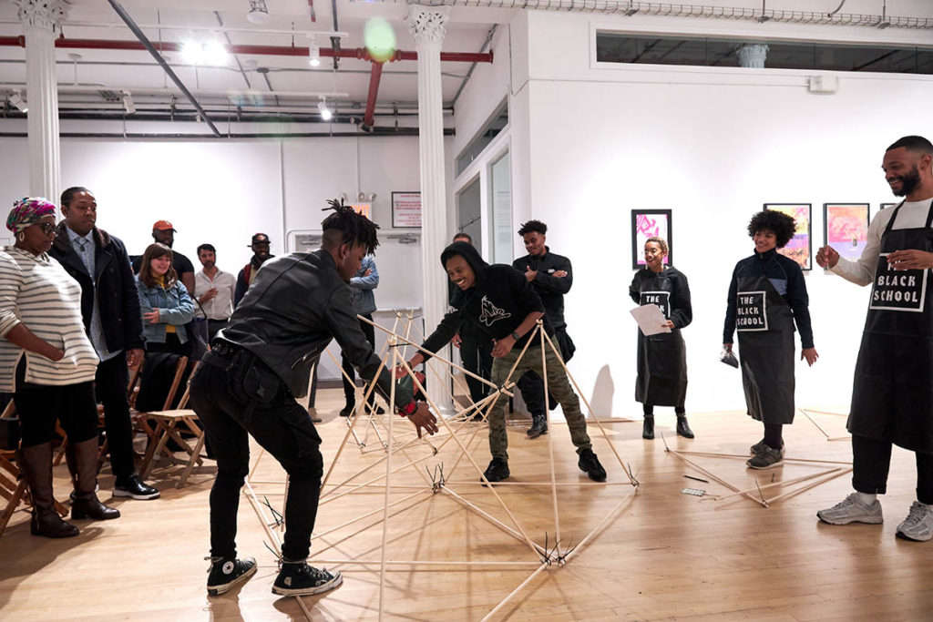 A group of people standing in a circle in a gallery space, with two people at the center working with a geometric structure made of wooden dowels.