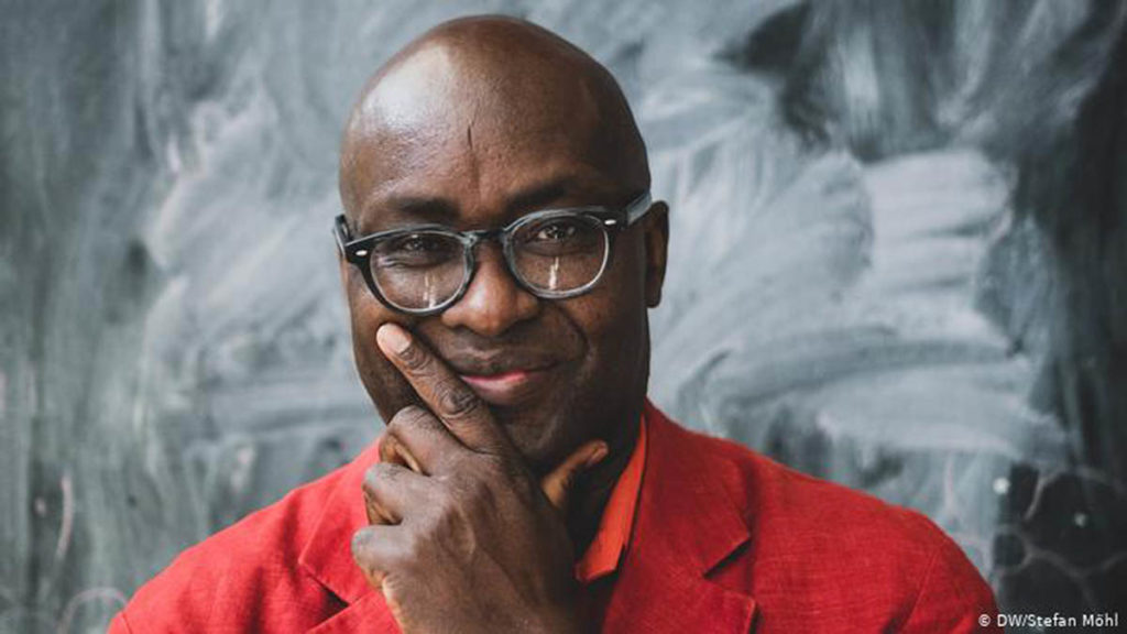 Headshot of Achille Mbembe, who wears a red shirt and glasses.