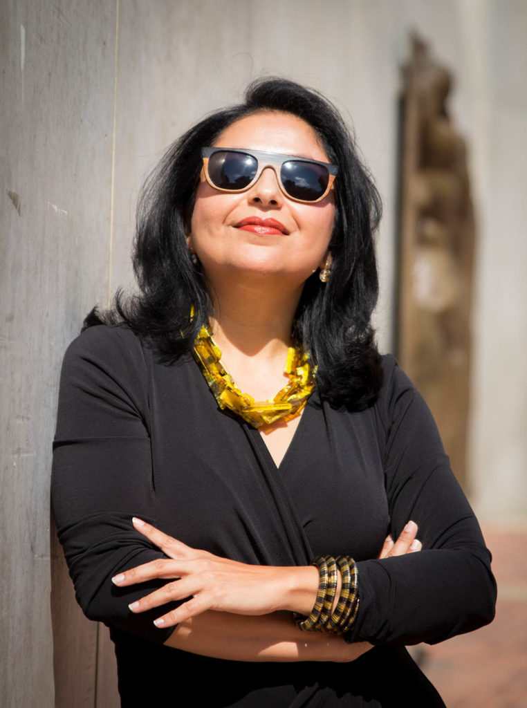 Headshot of Ananya Roy, who has long black hair and wears sunglasses, along with a large yellow necklace.