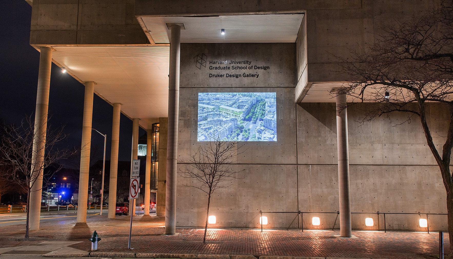 The Cambridge Street facade of Gund hall at night. On the wall is projected an image of a birds-eye view of a Paris block, including a rendering of a Dual-Use building.