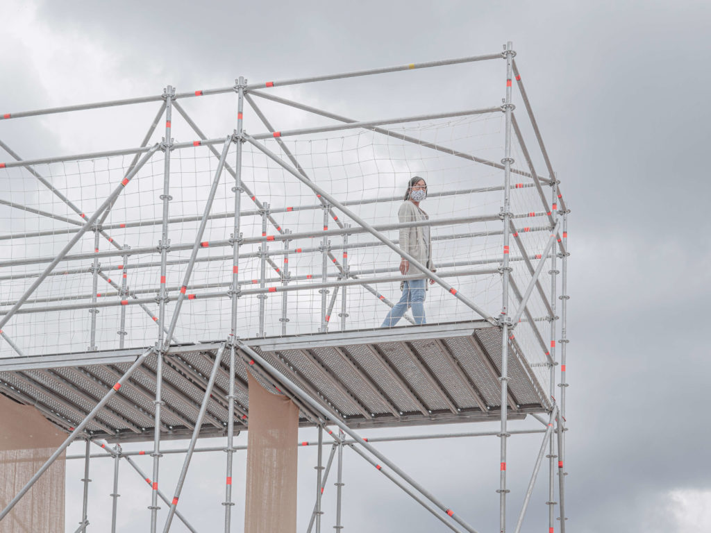 Masked woman walking on elevated scaffolding.