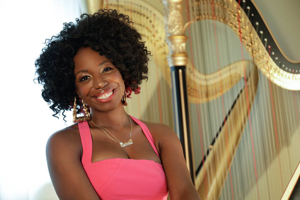 Headshot of Brandee Younger, who wears a pink top, has curly dark brown hair, and stands in front of a large gold harp.