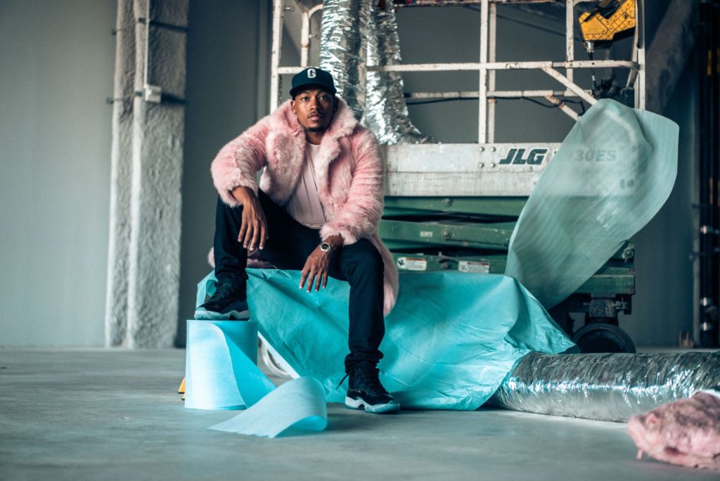 Portrait of Germaine Barnes sitting on brightly-colored construction material in front of a construction lift. Germain is wearing a baseball hat, pink fur coat, pink t-shirt, jeans and sneakers.