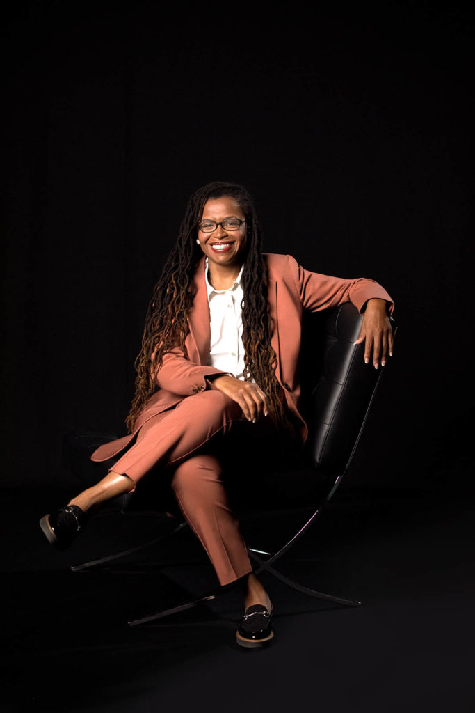 Photo of Dr. Andrea Roberts, who wears a salmon-colored suit with a white shirt and has long brown hair. She sits in a black chair in front of a black background.