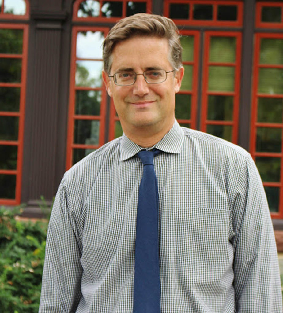 Headshot of Matthew Mulcahy who wears a collared shirt and blue tie, as well as glasses.