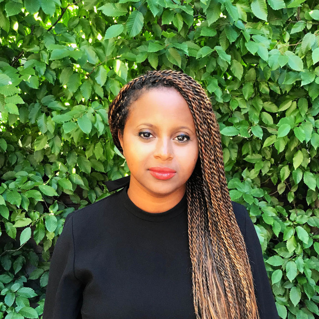 Headshot of Sara Zewde, who wears black and stands in front of a wall of leaves.