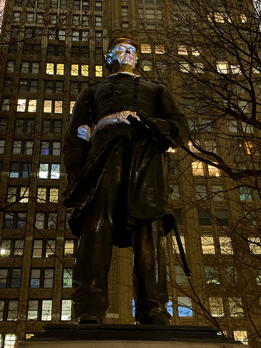 Photograph of a statue of a figure outdoors, in front of a tall building.