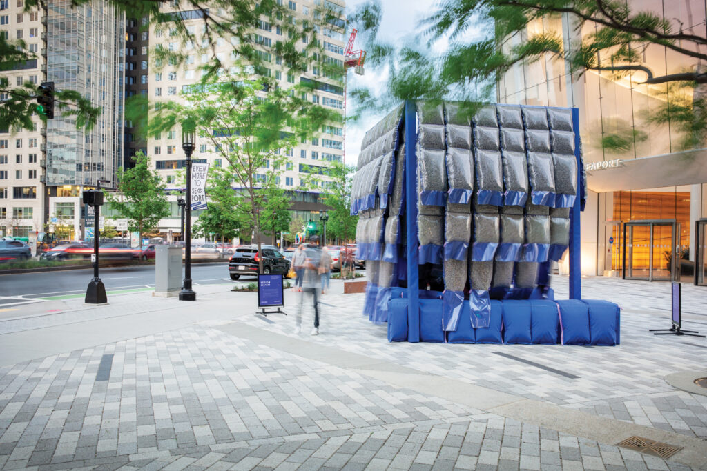 A pavilion sits in the centre of a plaza in Seaport, Boston. The pavilion is cobalt-blue and gray, with layers of puffy polished vinyl cells packed with recycled denim, hung from a timber frame in layers.