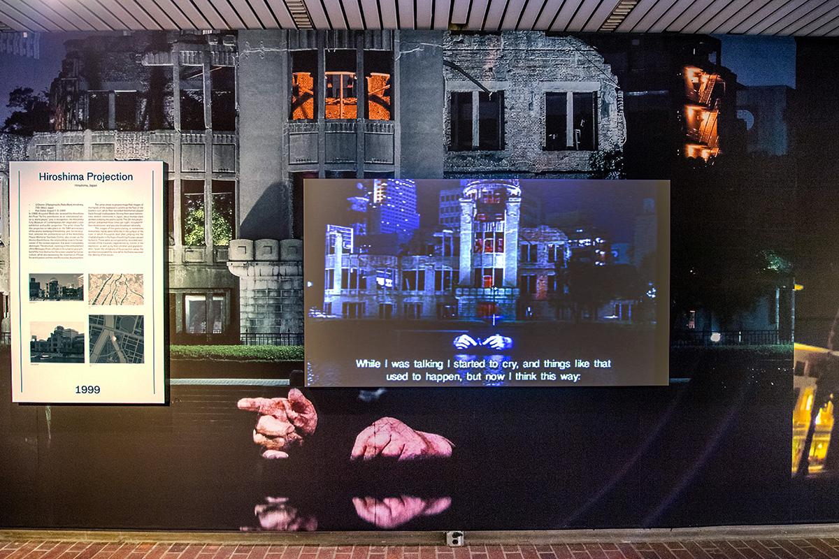 A wall in the lobby of Gund hall, displaying a text panel, mural, and video screen showing images of the Hiroshima Peace Memorial building with gesturing hands projected on the wall of the river bank below.
