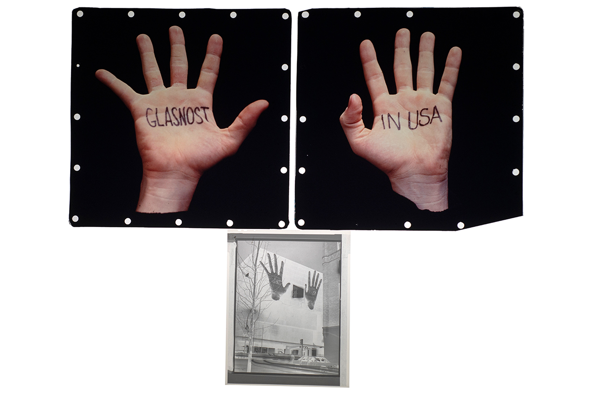 A detail of the light box showing an image of a person’s right hand with the word “GLASNOST” written on the palm, an image of a person’s left hand with the words “IN USA” written on the palm, and an image of those previous images projected on the side of a building.