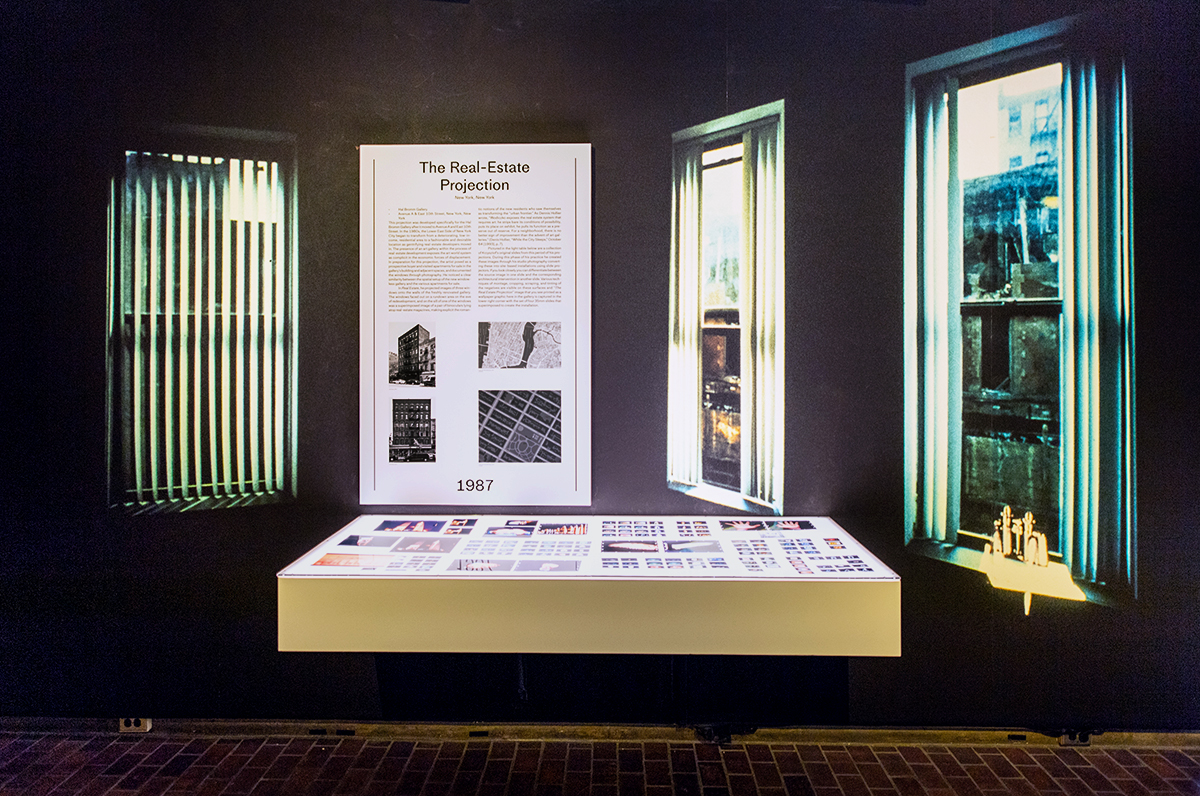 Photo of a gallery wall in Gund Hall showing a large image of Wodiczko’s Real Estate Projection, and a light table displaying a collection of slides.