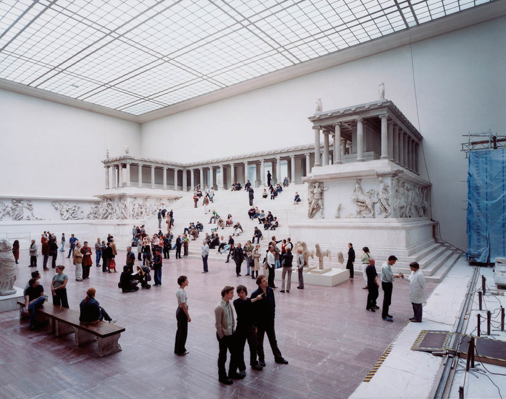 A large museum gallery with an architectural reproduction of a classical building. The gallery is filled with many people and has a roof with many windows.