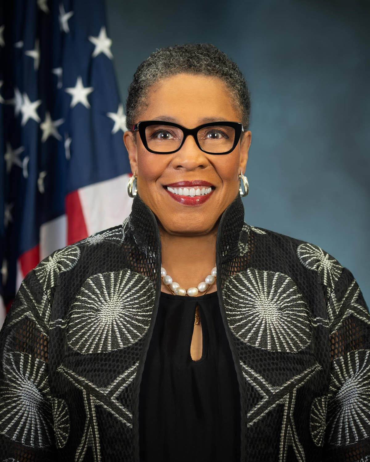Marcia L. Fudge stands in front of an American flag and a blue background, wearing black glasses.