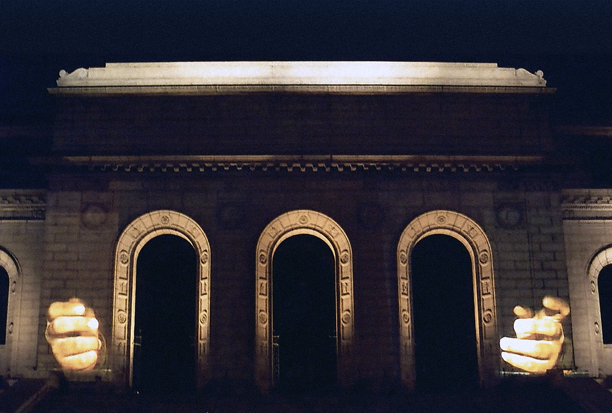 Photographic image of the facade of the Central Library at night, with three large arched doorways in the center. On either side of the arched doorways are video projections of human hands gesturing.