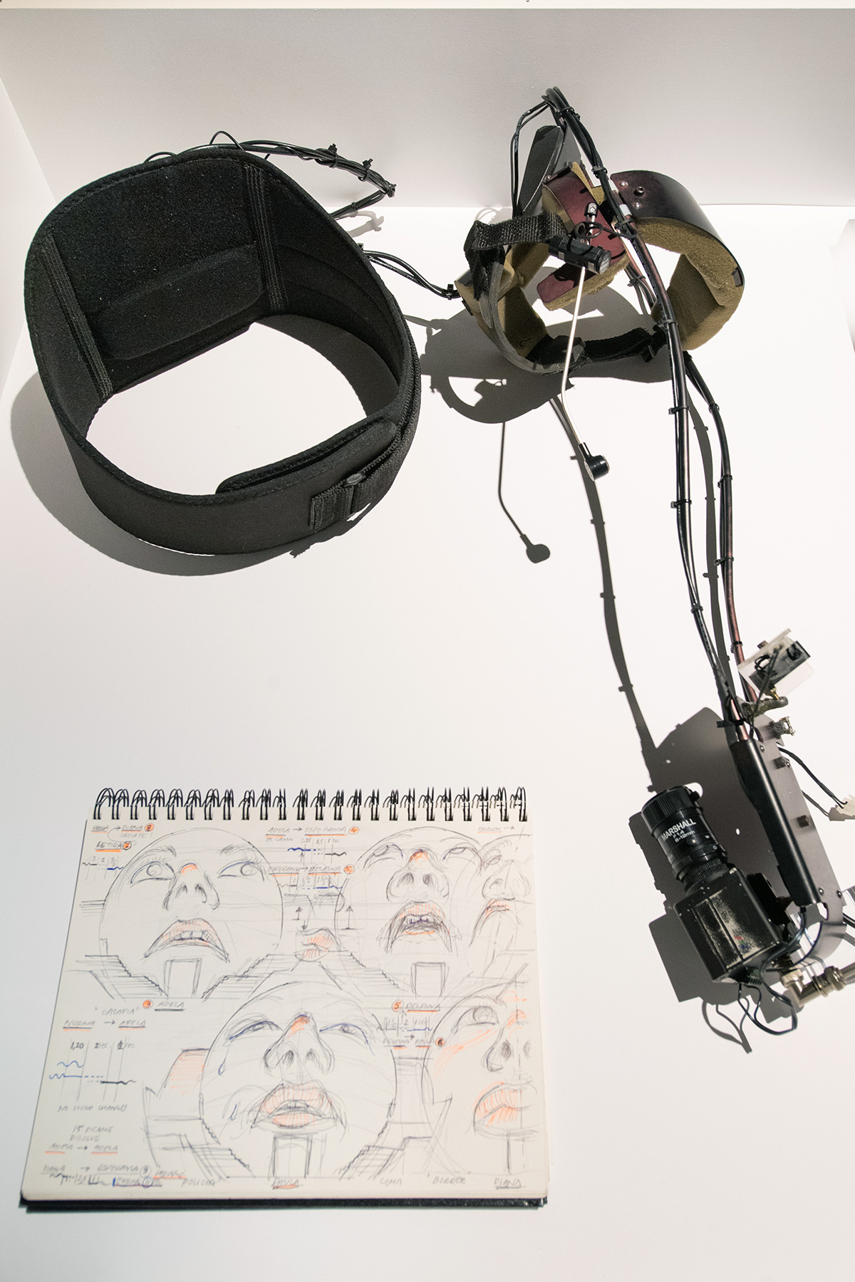 An apparatus made of metal and wire with electronics and a camera attached that is made to wear over a person’s head, and a sketchbook open to a page with pen and ink drawings of faces showing different expressions.