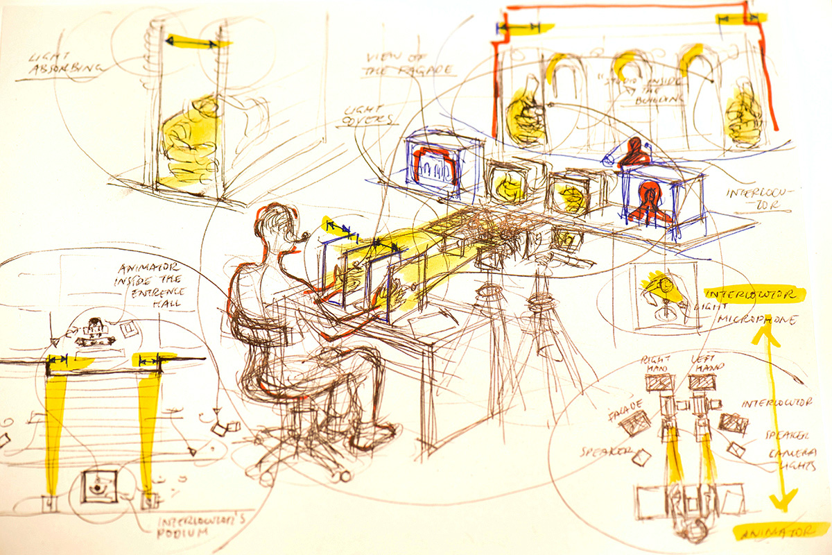 Pen and ink drawing in black and white with red and yellow highlights. The drawing depicts a person seated at a table with their hands in a boxed apparatus with video cameras pointed at the hands. Surrounding this drawing are additional sketches of hands and a building facade.