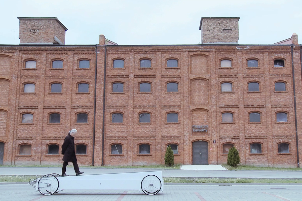 A man walks on a long low vehicle made of two long boxes with bicycle wheels and drivetrain attached, in front of a brick building.