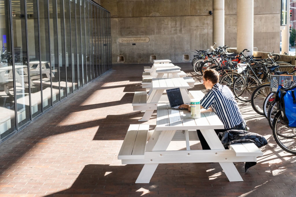 A photo of a student sitting at a white picnic table in the front plaza of Gund Hall that faces the street.