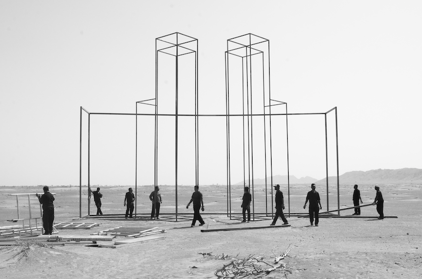 Figures walking around a desert landscape at the base of an abstract built structure.