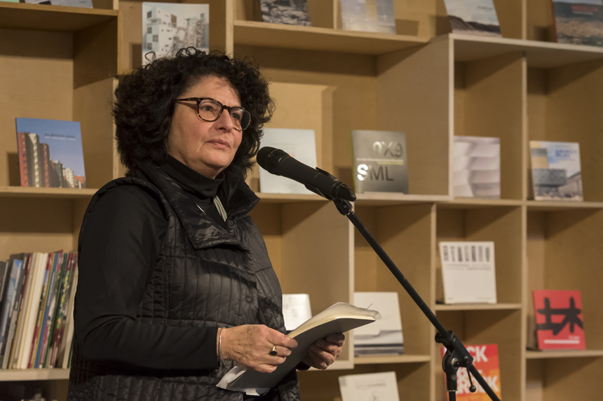 Portrait of Eve Blau speaking at microphone holding a book
