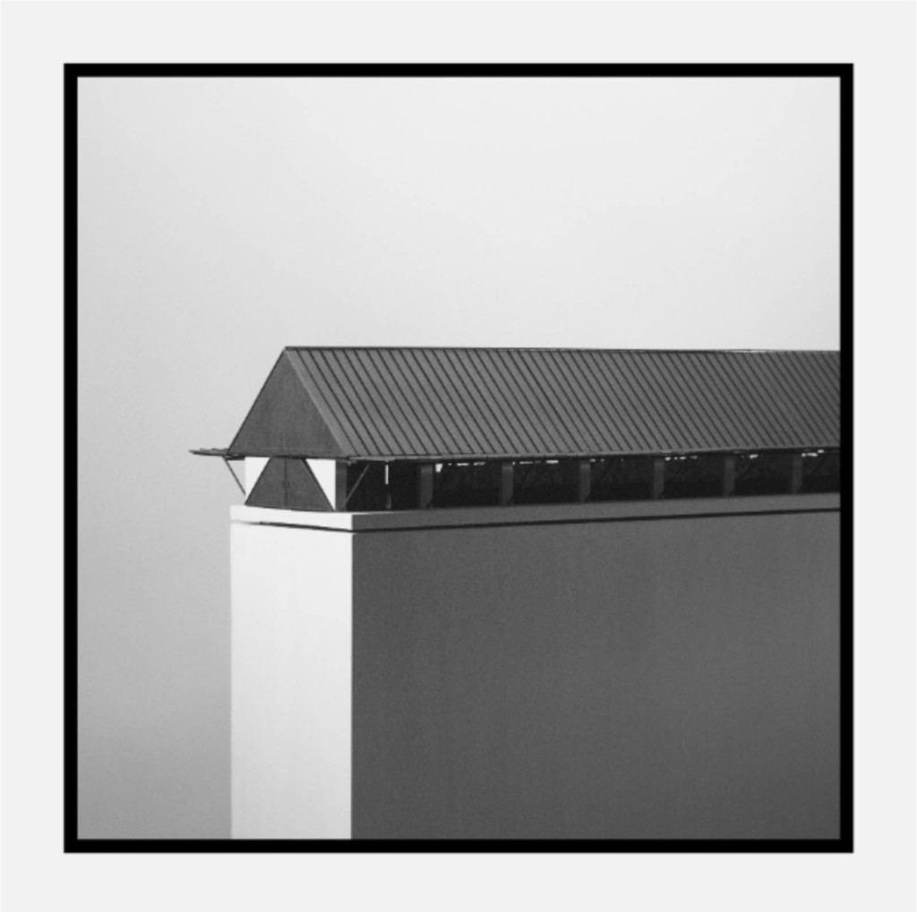 Black and white photo of wood architectural model shown on angle; structural is one story and long with a moderately sloped roof