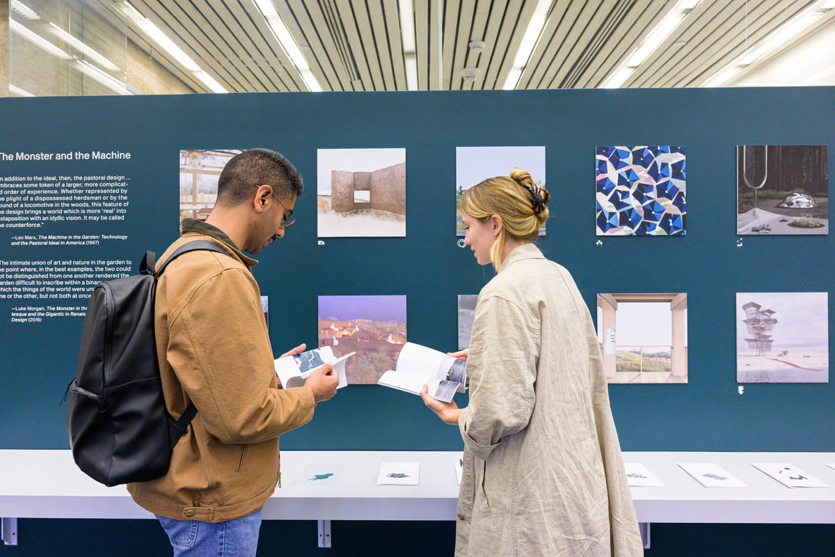 Two people look through exhibition pamphlets infront of the pamphlet display shelf and exhibit wall.