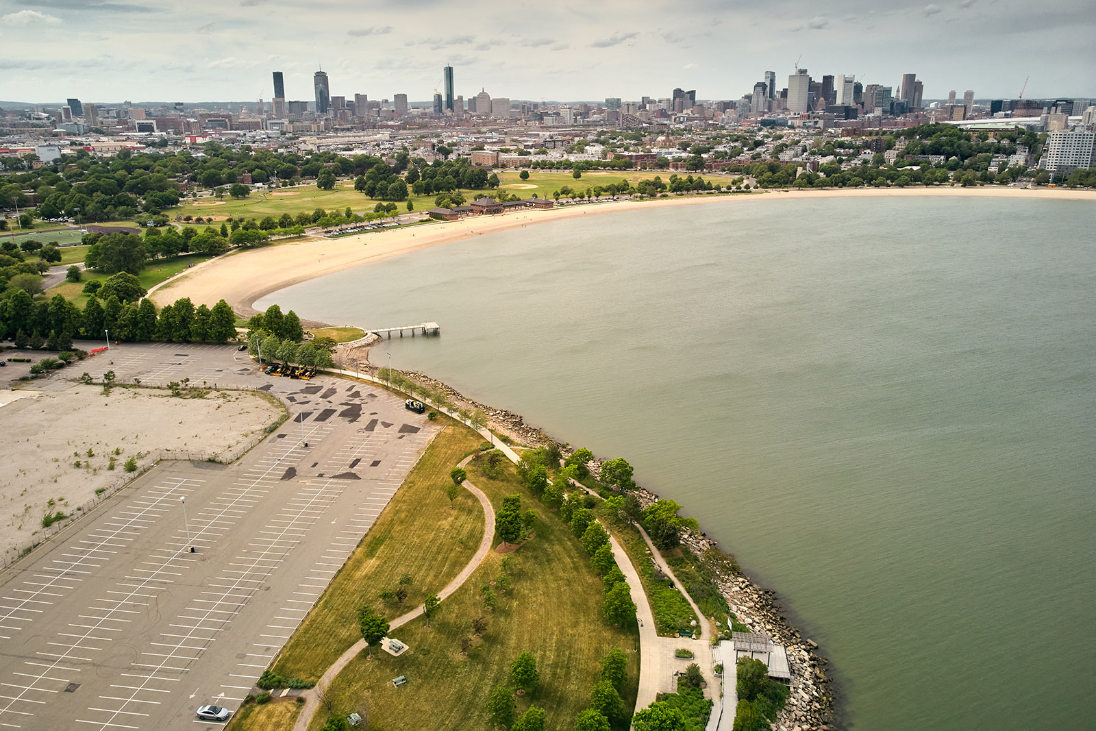 Aerial View of Dorchester Bay with empty parking lot on the left and ocean on the right. Beach and a skyline of Boston in a distance.