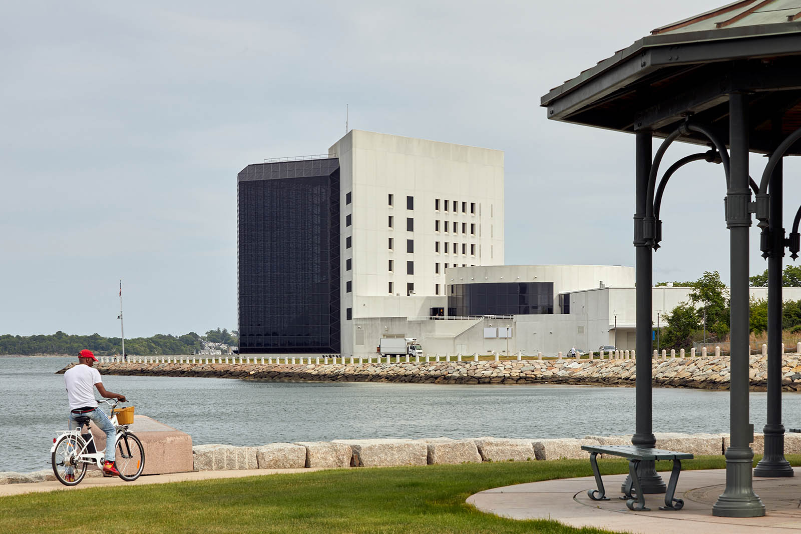 Photo of a half glass and have concrete building, a bay in front of it with a man on a bicycle looking at the building across the bay. A gazebo is shown on the right side of the photo