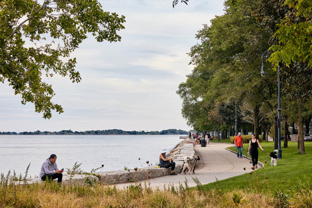 Photo of people walking along the water edge. On the left a man looking at his mobile phone, on the right side a woman walking a dog.
