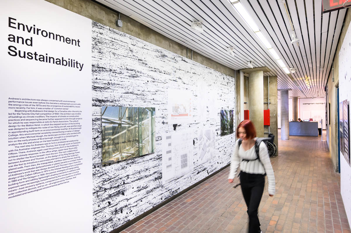 A person passing and exhibition wall diplaying a large text panel, new photographs, and old drawings; all against a stylized cement textured background.