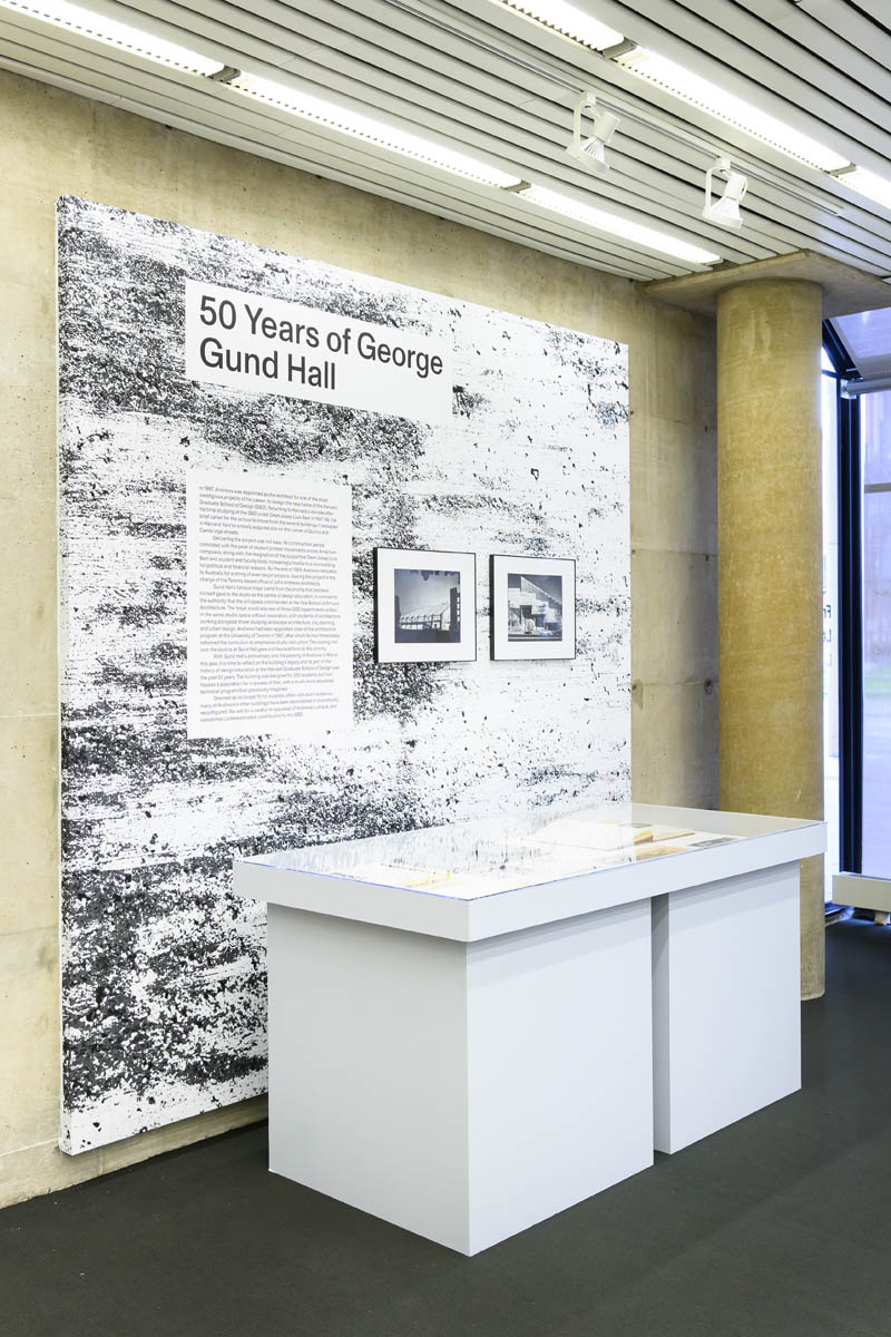 A display case in front of a wall displaying a title, small text panel, and two framed photographs of Gund Hall; all against a stylized cement background.