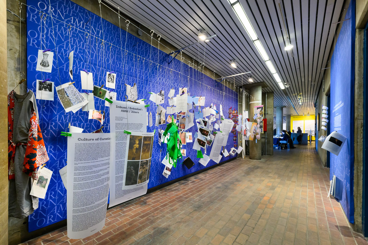 Blue exhibition Wall with a white text texture and printed images and text held onto wires with many colored clothes pins.