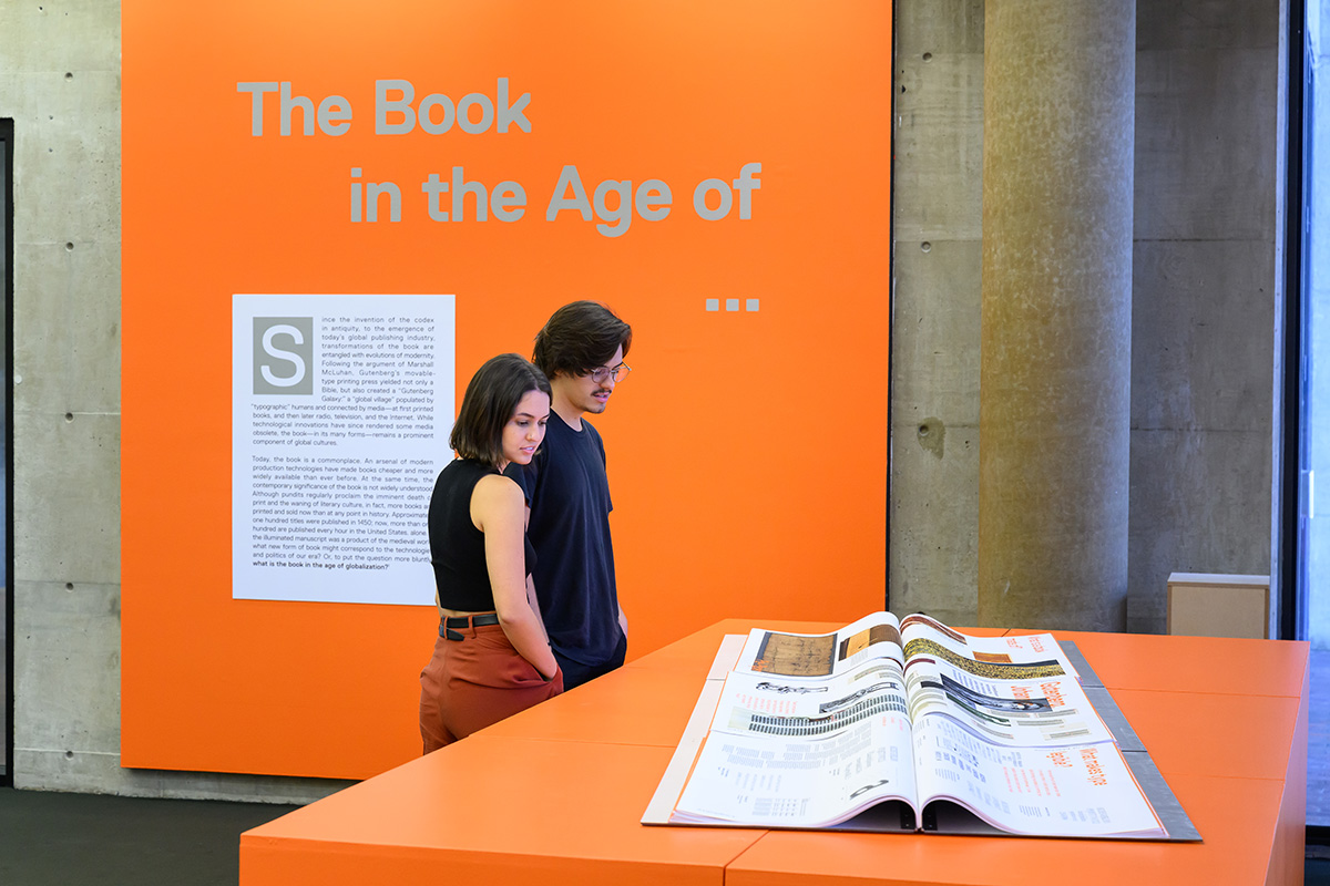 Two people looking at a large book on display that spans several tables in Frances Loeb Library.